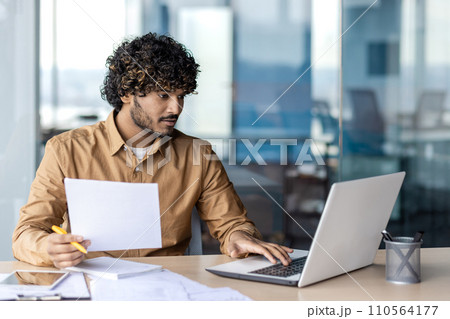 Engaged young adult with curly hair reviews documents while using laptop in a bright, contemporary office space, showcasing productivity. Engaged young adult with curly hair reviews documents while using laptop in a bright, contemporary office space, showcasing productivity. 110564177