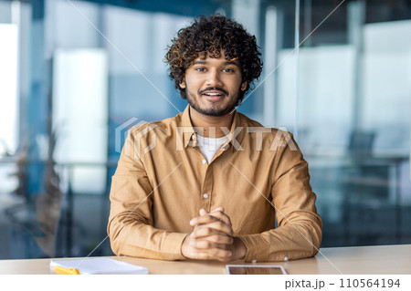A professional young businessman with curly hair sits confidently at a desk in a contemporary, well-lit office environment, smiling at the camera. 110564194