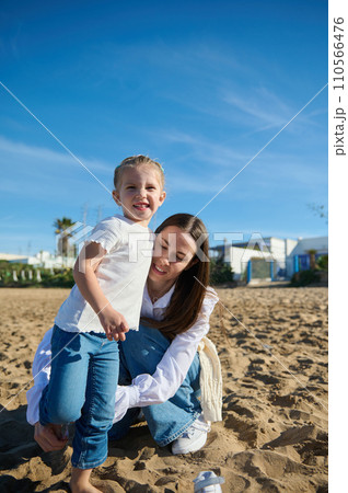 Adorable little child girl smiling looking at the camera, playing with her mom on the sandy beach on warm sunny day Adorable little child girl smiling looking at the camera, playing with her mom on the sandy beach on warm sunny day 110566476