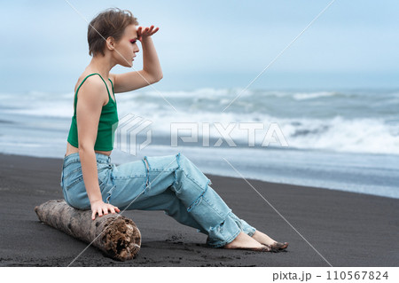 Hipster woman sitting on log on black sandy beach of ocean, raised hand to forehead, looking at view Hipster woman sitting on log on black sandy beach of ocean, raised hand to forehead, looking at view 110567824