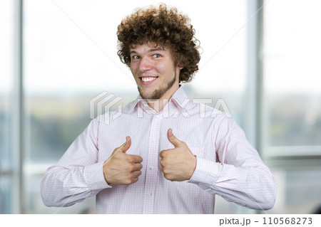 Happy smiling young man with curly hair shows both thumbs up. Indoor window in the background. Happy smiling young man with curly hair shows both thumbs up. Indoor window in the background. 110568273