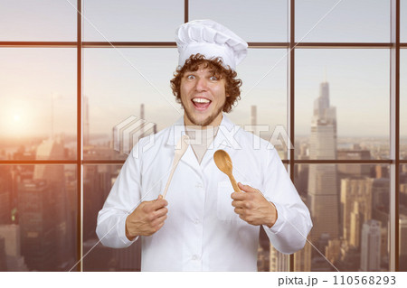 Portrait of a happy young caucasian male cook with a wooden spoon. Checkered window with cityscape in the background. Portrait of a happy young caucasian male cook with a wooden spoon. Checkered window with cityscape in the background. 110568293