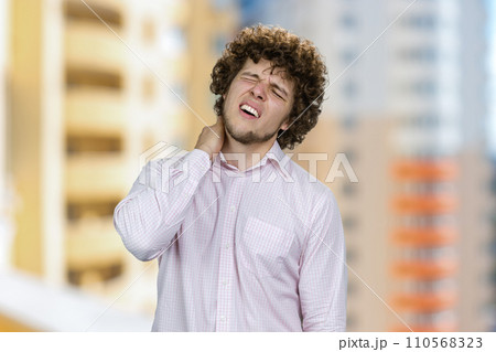 Young handsome man with curly hair having neckache. Residential area in the background. 110568323