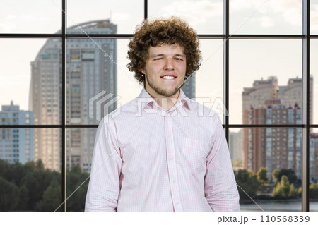 Happy young attractive man with curly hair bites his lip. Indoor window in the background. Happy young attractive man with curly hair bites his lip. Indoor window in the background. 110568339