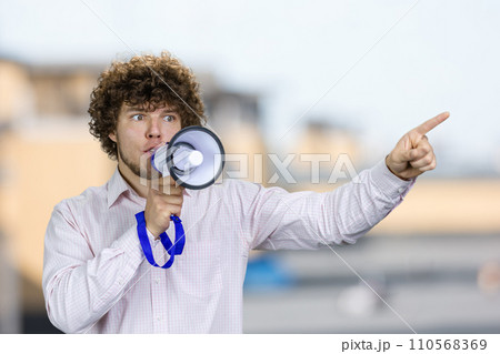 Young man with curly hair in white shirt speaks in megaphone pointing with index finger. Blurred urban scape in the background. 110568369