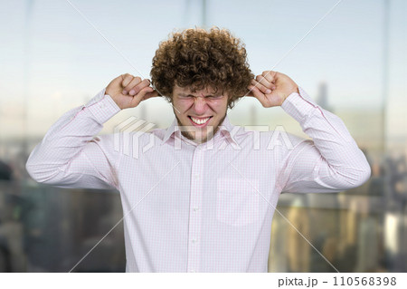 Caucasian young man in white shirt close his ears with both fingers. He doesn't want to hear that. Nervous breakdown concept. Blurred cityscape in the background. 110568398