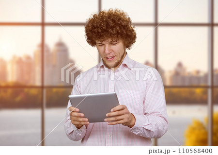 Portrait of a young caucasian man with curly hair playing a video game on the tablet pc device. Indoor window in the background. Portrait of a young caucasian man with curly hair playing a video game on the tablet pc device. Indoor window in the background. 110568400