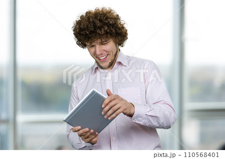 Portrait of a young happy caucasian man with curly hair playing a video game on the tablet pc device. Indoor window in the background. Portrait of a young happy caucasian man with curly hair playing a video game on the tablet pc device. Indoor window in the background. 110568401