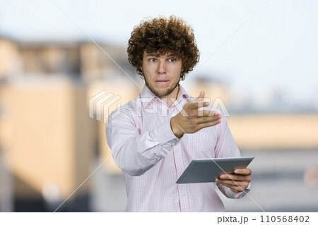 Portrait of a young caucasian worker with curly hair standing with tablet pc device pointing at something. Blurred urban scape in the background. Portrait of a young caucasian worker with curly hair standing with tablet pc device pointing at something. Blurred urban scape in the background. 110568402