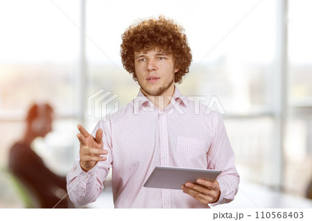Portrait of a young businessman with curly hair standing with tablet pc device pointing at something. Office environment in the background. 110568403