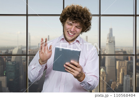Portrait of a young happy caucasian man holding the tablet device and waving his hand. Checkered window with cityscape in the background. 110568404