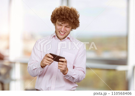Excited happy young caucasian male gamer playing using smartphone. Guy playing mobile videogame app or watching digital video stream holding cell phone indoors. Excited happy young caucasian male gamer playing using smartphone. Guy playing mobile videogame app or watching digital video stream holding cell phone indoors. 110568406