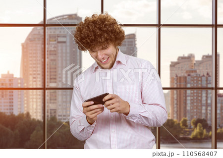 Excited happy young caucasian man gamer playing using smartphone. Guy playing mobile videogame app or watching a video stream holding cell phone indoors. Excited happy young caucasian man gamer playing using smartphone. Guy playing mobile videogame app or watching a video stream holding cell phone indoors. 110568407