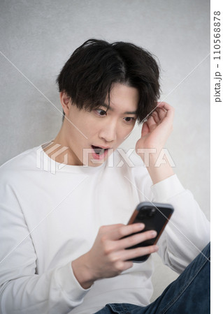 壁に寄りかかって座り笑顔でスマホを見て驚く男性のバストアップ 壁に寄りかかって座り笑顔でスマホを見て驚く男性のバストアップ 110568878