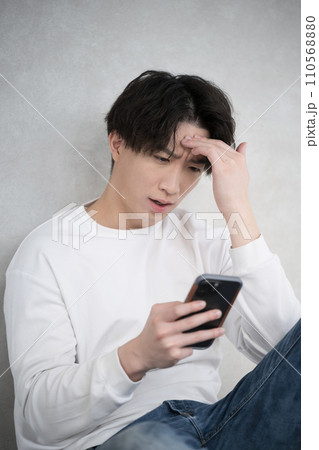 壁に寄りかかって座り笑顔でスマホを見て悩み頭を抱える男性の上半身 110568880