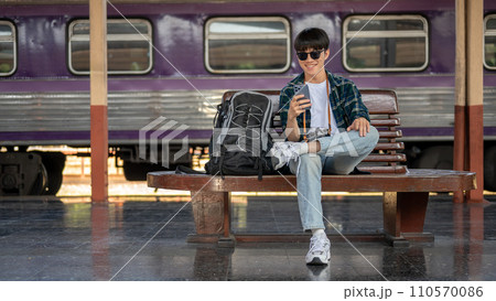 A young Asian male traveler is using his smartphone while waiting for his train at a railway station A young Asian male traveler is using his smartphone while waiting for his train at a railway station 110570086