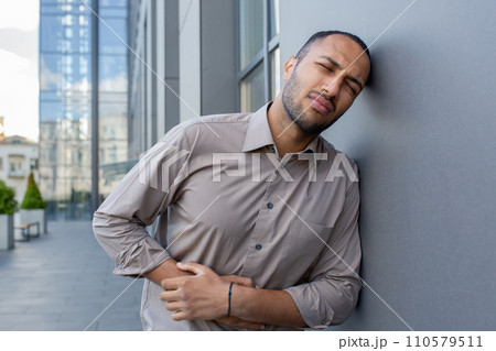Man in pain outdoors, clutching his side and grimacing, conveying a sense of discomfort or abdominal ache against an urban backdrop. Man in pain outdoors, clutching his side and grimacing, conveying a sense of discomfort or abdominal ache against an urban backdrop. 110579511