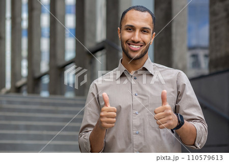 Confident young man in a casual shirt giving thumbs up with a smile, outdoor steps in the background, expressing success and positivity. 110579613