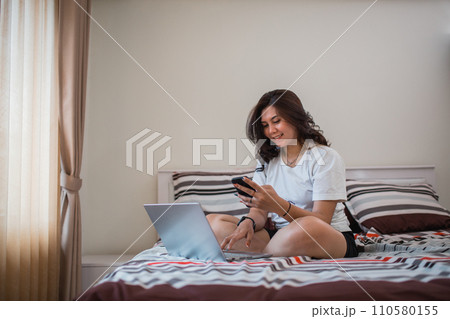 young Asian woman looking at cell phone while using laptop on bed 110580155