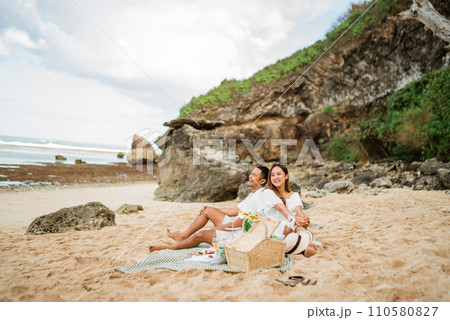 young Asian couple sitting jokingly on a picnic blanket on a beach 110580827