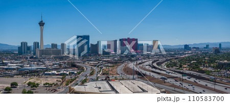 Aerial view of Las Vegas skyline featuring the Stratosphere Tower, iconic hotels, and mountains, under a clear blue sky with busy highways, capturing the city's dynamic essence. 110583700