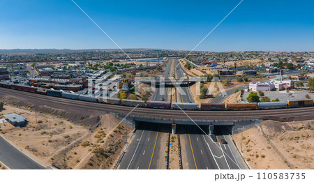 Aerial view of a southwestern American town with a freight train crossing a highway, surrounded by desert terrain, commercial and residential buildings under a clear blue sky. 110583735