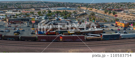 Aerial view of Barstow, California, showcasing Barstow Station, businesses, and vehicles amidst a sunny, semi-arid landscape with hills, clear skies, and scattered greenery. 110583754