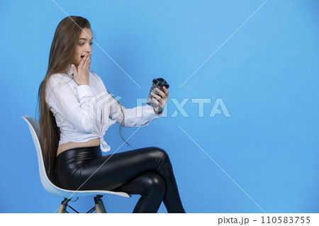 Tired young woman in elegant outfit, yawning, while drinking coffee in cozy white chair. Side view of graceful lady with coffee cup, covering mouth with palm, isolated on blue. Concept of cheering up. Tired young woman in elegant outfit, yawning, while drinking coffee in cozy white chair. Side view of graceful lady with coffee cup, covering mouth with palm, isolated on blue. Concept of cheering up. 110583755