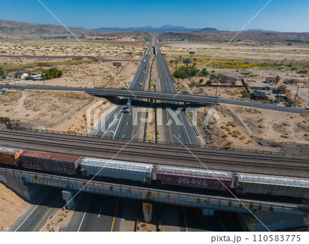 Aerial view of a sunlit highway interchange and parallel train tracks in a desert landscape. Sparse vegetation and distant rugged mountains under a clear blue sky. 110583775