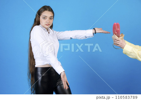 Excited long-haired woman shrugging, outstretching hand, while gesturing at wrapped pot of prickly cactus plant, presented by anonymous crop hand, isolated on blue background. Concept of gesturing. 110583789
