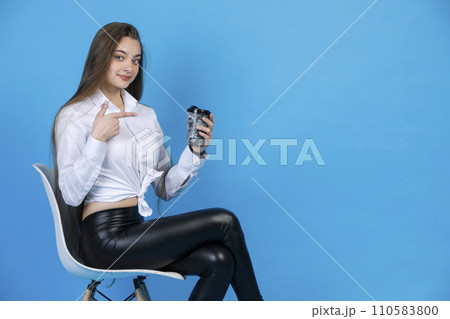 Smiling charming girl in black leggings, pointing at cup of tasty coffee, while sitting in chair. Side view of graceful lady gesturing, while posing, isolated on blue background. Concept of gesturing. Smiling charming girl in black leggings, pointing at cup of tasty coffee, while sitting in chair. Side view of graceful lady gesturing, while posing, isolated on blue background. Concept of gesturing. 110583800