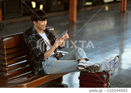 Happy Asian male traveler with suitcase using mobile phone while waiting train. Travel and vacations concept. Happy Asian male traveler with suitcase using mobile phone while waiting train. Travel and vacations concept. 110584005
