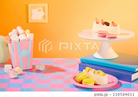 A table of sweets. Marshmallow inside a paper box, cakes, cookies and macarons are displayed on a plate. Orange background with checkered surface. Free space for display. A table of sweets. Marshmallow inside a paper box, cakes, cookies and macarons are displayed on a plate. Orange background with checkered surface. Free space for display. 110584011
