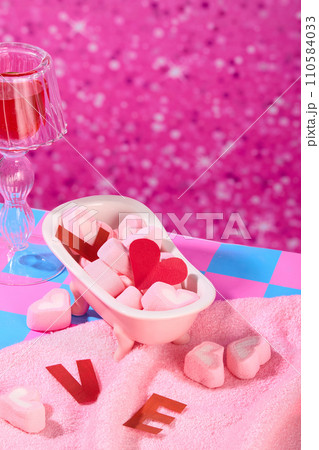 Heart-shaped marshmallow is contained inside a mini bathtub, a pink towel, next to a decorative glass on a sparkling pink background. Copy space with close up. 110584033