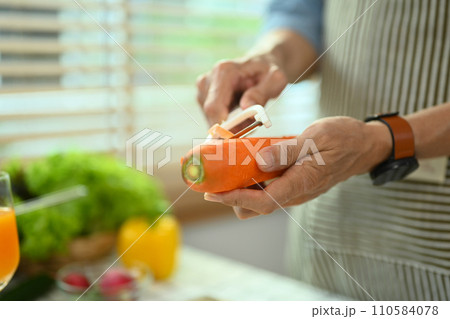 Cropped shot senior man in apron peeling carrot, preparing healthy salad in kitchen Cropped shot senior man in apron peeling carrot, preparing healthy salad in kitchen 110584078
