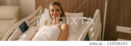 Smiling woman lying on bed at modern hospital ward after surgery. Health care and medicine concept Smiling woman lying on bed at modern hospital ward after surgery. Health care and medicine concept 110586191