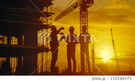日の出時、建設現場で設計図を議論する建設チーム、夜明けの空に映えるシルエット。 110586473