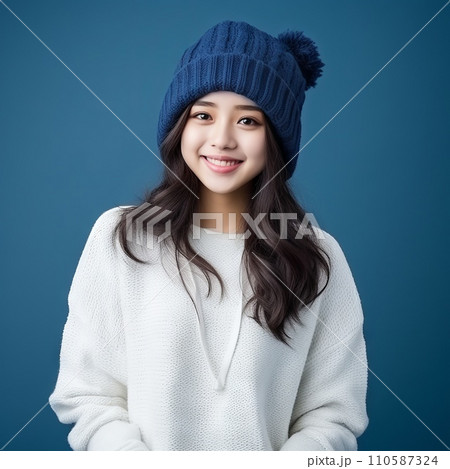 Happy asian teenage girl in a blue winter hat and white sweater. Portrait of a teenager girl in a winter cap smiling on a blue background looking at the camera. Cheerful Japanese teen in pom pom hat. 110587324