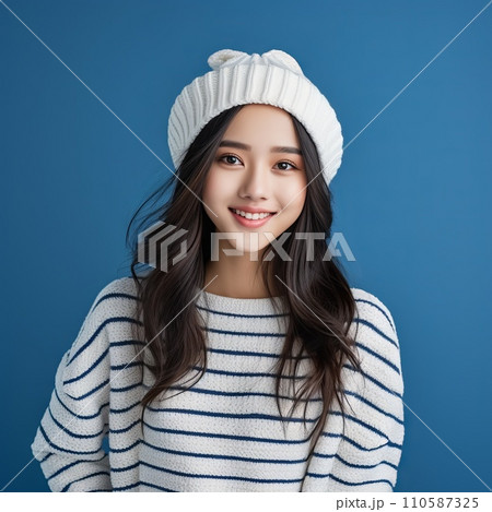 Happy asian teenage girl in a white winter hat and white sweater. Portrait of a teenager girl in a winter cap smiling on a blue background looking at the camera. Cheerful Japanese teen in pom pom hat. 110587325