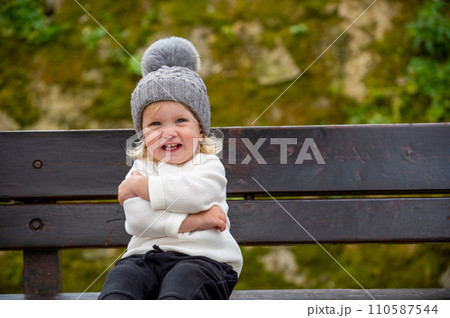 Cute blonde kid sitting on a bench outside in a park, smiling. Happy child wearing a hat outdoors on a spring day. Adorable grinning caucasian boy resting on a bench after play time on a playground. 110587544