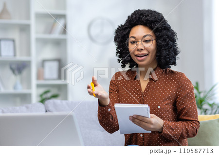 Young African American female teacher sitting on the sofa at home, holding a notebook and teaching remotely via laptop, explaining and gesturing to the camera. Young African American female teacher sitting on the sofa at home, holding a notebook and teaching remotely via laptop, explaining and gesturing to the camera. 110587713
