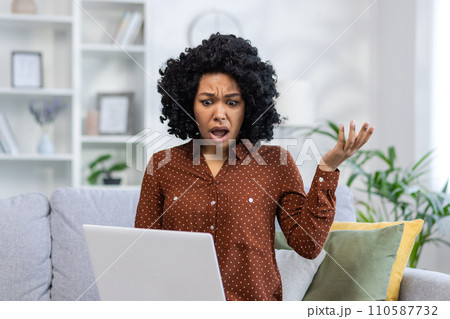 Angry young African American woman sitting on the couch at home and looking at the laptop screen with her hands in frustration. 110587732