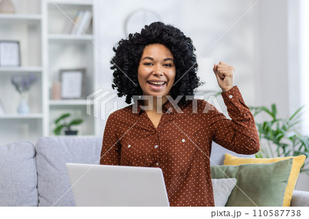 Portrait of a happy African American woman sitting at home on the couch in front of the camera and celebrating her success by raising her hand up 110587738