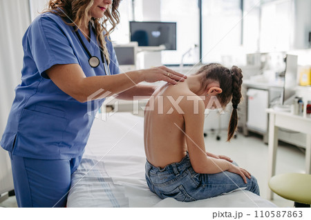 Orthopedist examines the spine, posture, and spinal deformities of little girl. Girl visiting paediatrician for annual preventive exam. Concept of preventive health care for children. 110587863