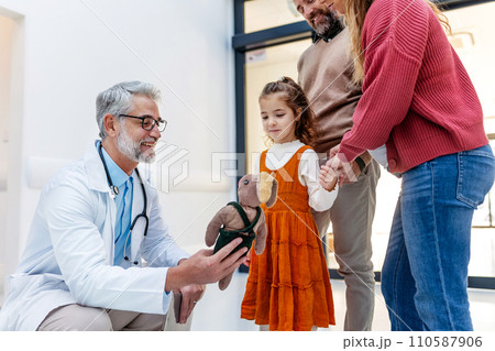 Pediatrician looking at girl's toy. Girl patient is at examination in a modern clinic with her parents. Emotional support during medical examination. 110587906