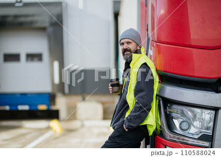 Truck driver leaning against red truck and drinking coffee, waiting for warehouse workers. 110588024
