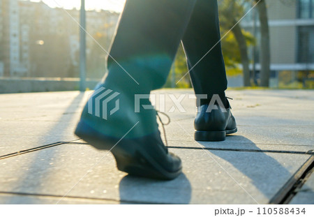 Legs of a businessman in fashionable shoes walking outdoors. Business concept. Close-up view to the businessman in a black new shoes walks on the street. Stylish men wears. Low angle. Rear view. Legs of a businessman in fashionable shoes walking outdoors. Business concept. Close-up view to the businessman in a black new shoes walks on the street. Stylish men wears. Low angle. Rear view. 110588434