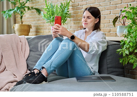 Young happy woman using smartphone, sitting on sofa at home 110588751
