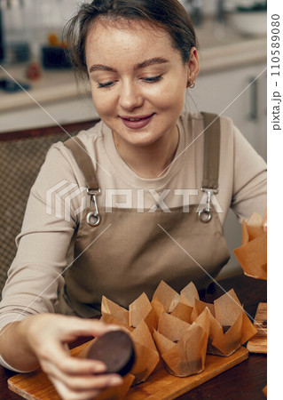 A young woman packs cupcakes in a craft box. the concept of food delivery. A birthday package. small business. The bakery chef bakes pastries and cakes in the kitchen A young woman packs cupcakes in a craft box. the concept of food delivery. A birthday package. small business. The bakery chef bakes pastries and cakes in the kitchen 110589080