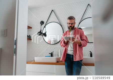 Man shopping online while brushing his teeth in bathroom. Holding tablet, scrolling and buying in eshop, toothbrush in mouth. 110589549